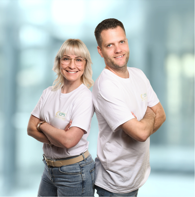 Caro und Pascal stehen lächelnd Rücken an Rücken und blicken direkt in die Kamera.